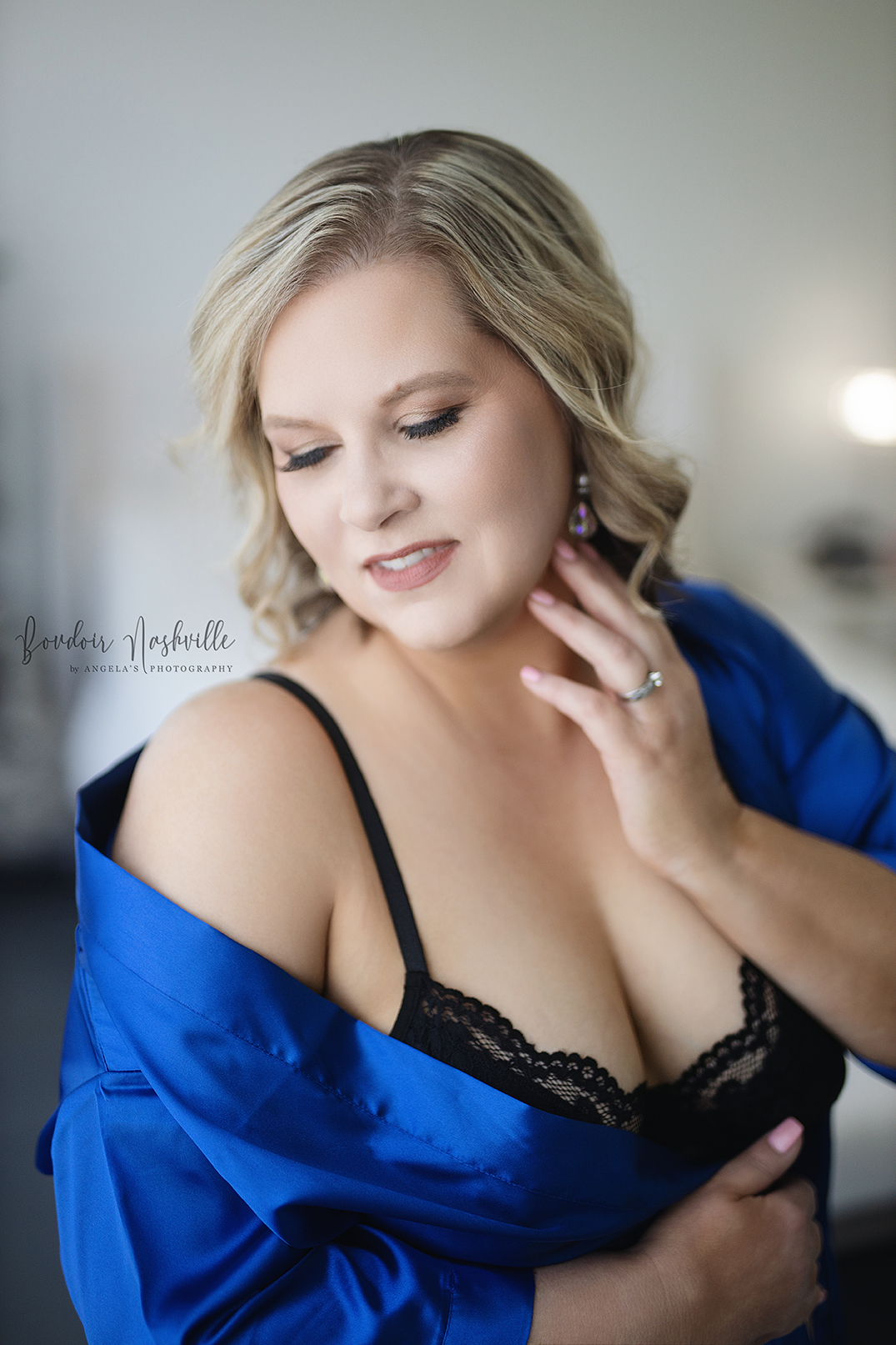 Confident woman with blonde hair wearing a vibrant royal blue satin robe draped off her shoulders, revealing a delicate black lace bralette. She is softly smiling with her eyes closed, exuding a sense of self-love and empowerment. Her hand gently touches her neck, highlighting elegant earrings and a simple silver ring. The background is softly blurred, adding to the intimate and dreamy atmosphere.